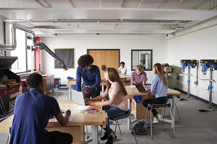teacher-talking-female-high-school-student-sitting-work-bench-using-laptop-design-technology-lesson
